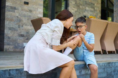 Photo for Woman blows on the wound on her sons knee, they are located on the terrace of a country house - Royalty Free Image