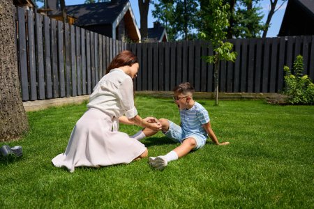 Téléchargez les photos : Mère souriante console son fils sur la pelouse verte, le garçon s'est blessé au genou et pleure - en image libre de droit
