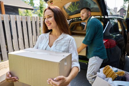 Photo for Beautiful woman carries a box of things from the trunk, she and her husband unload the car - Royalty Free Image