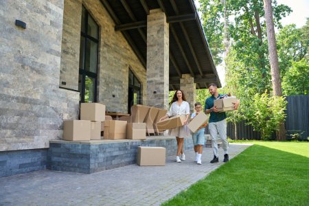 Photo for Parents and their son are moving to beautiful new house, they are carrying a lot of cardboard boxes with things - Royalty Free Image