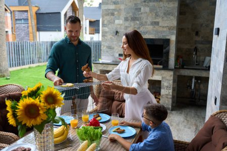 Téléchargez les photos : Femme met de la nourriture chaude barbecue sur les assiettes, tournesols et fruits sur la table - en image libre de droit