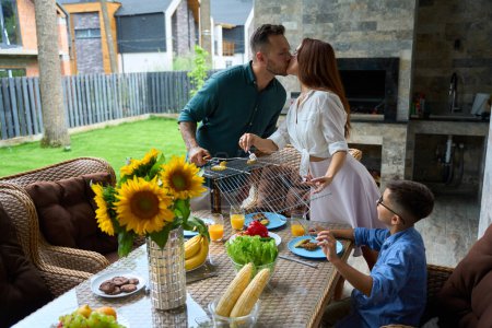 Photo for Family gathered for lunch in the barbecue area, parents put food on plates and kiss - Royalty Free Image