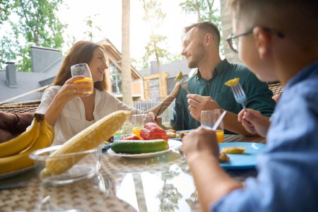 Foto de Mamá, papá e hijo están hablando durante el almuerzo en la terraza, hay comida deliciosa y jugo en la mesa - Imagen libre de derechos