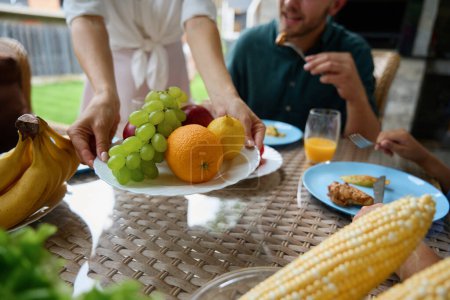Téléchargez les photos : Femme sert des fruits sur un plateau, une famille est assise sur la terrasse à la table à manger - en image libre de droit