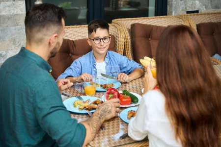 Foto de Los padres se comunican con su hijo durante el almuerzo en la terraza, hay un montón de deliciosa comida en la mesa - Imagen libre de derechos
