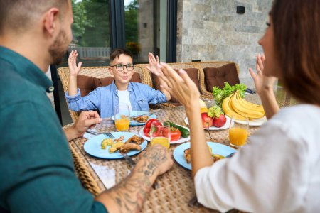Foto de Madre y padre se comunican con su hijo durante el almuerzo en la terraza, la gente usa muebles de jardín cómodos - Imagen libre de derechos