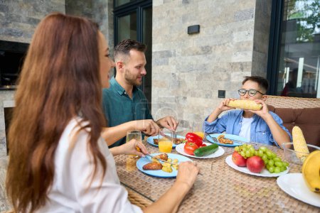 Téléchargez les photos : Adolescent mange du maïs avec appétit, une famille déjeune sur la terrasse d'une maison de campagne - en image libre de droit