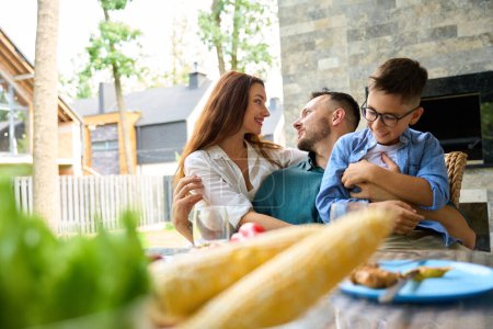 Photo for Man tenderly hugs his wife and son, the family sits on the terrace at the dining table - Royalty Free Image