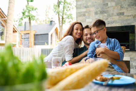 Photo for Woman tenderly hugs her husband and son, the family sits on the terrace at the dining table - Royalty Free Image
