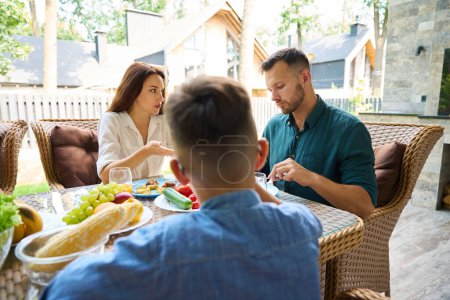 Téléchargez les photos : Jeune couple se disputent au déjeuner sur la terrasse, leur fils adolescent est présent à la table - en image libre de droit