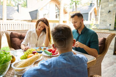 Foto de Hombre y una mujer están sentados molestos en el almuerzo en la terraza, su hijo adolescente está presente en la mesa. - Imagen libre de derechos