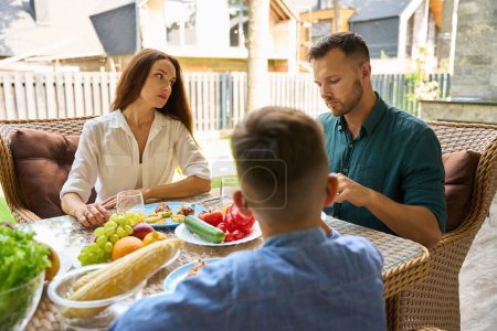 Foto de Mujer y un hombre están sentados molestos en el almuerzo en la terraza, su hijo adolescente está presente en la mesa - Imagen libre de derechos