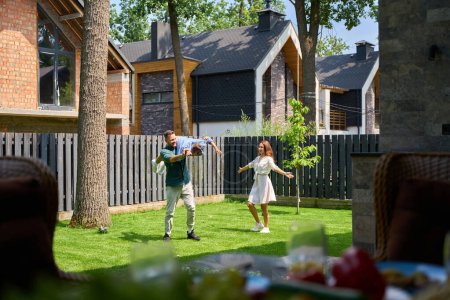 Photo for Joyful mother and father are having fun playing with their son in front of a country house, they are happy - Royalty Free Image