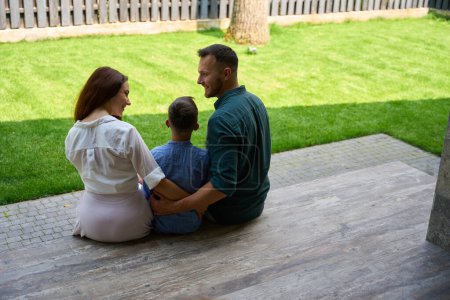 Photo for Happy family sits on the terrace of a country house, parents tenderly hug their son - Royalty Free Image