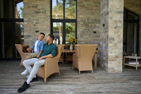 Foto de El hombre y su hijo adolescente se comunican en la terraza de una casa de campo, que están sentados en sillas de jardín - Imagen libre de derechos