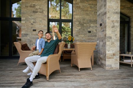 Photo for Male and his teenage son are talking on the terrace of a country house, they are seated in garden chairs - Royalty Free Image