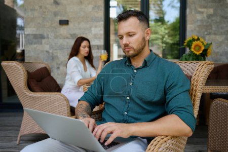 Photo for Man sits in a wicker chair and works on a laptop, in the distance his wife sits at the table - Royalty Free Image