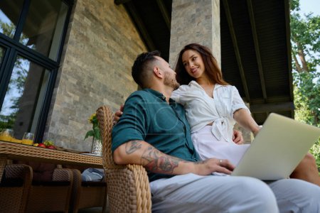 Photo for Male in a wicker chair and working on a laptop, his wife gently hugs him - Royalty Free Image