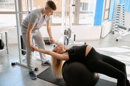 Photo for Man instructor works out with a pregnant woman in the gym of a health center, using a fitball - Royalty Free Image