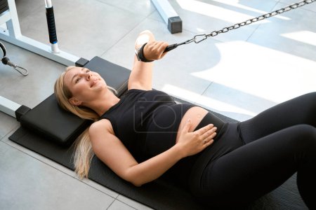 Foto de Mujer sonriente en ropa deportiva está haciendo ejercicio mientras está acostada en una máquina de ejercicio especial, la mujer está embarazada - Imagen libre de derechos