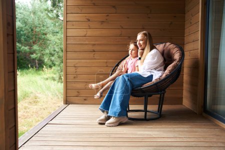 Foto de Sonriente hija y atractiva madre sentada en un sillón en un gran porche y admirando la naturaleza, respirando aire fresco del bosque, pasatiempo juntos - Imagen libre de derechos