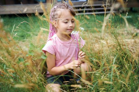Foto de Chica bonita con peinado elegante sentado en la hierba y la recogida de pequeñas flores silvestres en ramo, disfrutando de un clima cálido de verano y respirar aire fresco - Imagen libre de derechos