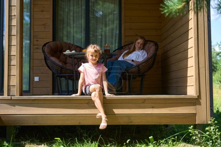Photo for Attractive mother sitting in cozy garden armchair on wooden porch of country house and watching her little daughter playing nearby in grass, enjoying fresh air - Royalty Free Image
