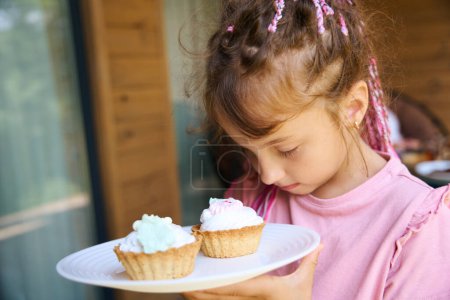 Photo for Close-up little pretty girl looking at sweet creamy cakes lying at plate with appetite, wants to eat them on lunch, sweet-tooth baby - Royalty Free Image