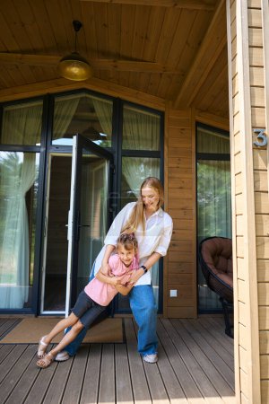 Téléchargez les photos : Mère et fille s'amusent à passer du temps en plein air, fille penchée d'un côté à l'autre et mère attrapant son debout sur le porche en bois de la maison de campagne familiale - en image libre de droit