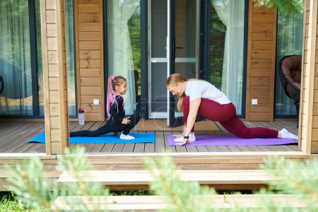 Photo for Active mother and daughter doing sport on wooden porch of their house, doing exercises and stretching body standing on yoga mats, practicing yoga at home - Royalty Free Image