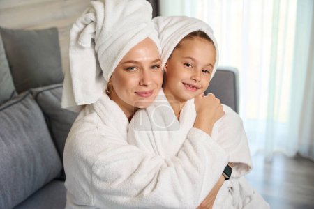 Téléchargez les photos : Belle mère et jolie petite fille en peignoirs doux blancs et serviettes de bain sur les têtes étreignant et posant en regardant la caméra - en image libre de droit