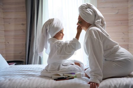 Photo for Cute little girl in big bathrobe and with towel on her head applying make-up on pretty clean face of her mother, girls having fun after spa together - Royalty Free Image