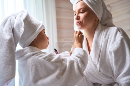 Foto de Close-up bonita hija pintando los labios madre con brillo labial después del procedimiento de spa, chicas pasar tiempo juntos en albornoces - Imagen libre de derechos