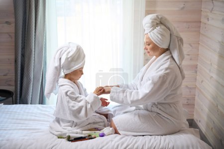 Téléchargez les photos : Fille mignonne appliquant crème de douceur sur les mains de la mère, procédure de beauté après spa, passe-temps de plaisir ensemble dans les peignoirs et les serviettes - en image libre de droit
