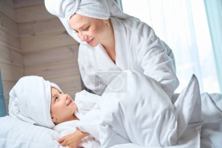 Téléchargez les photos : Mère trouvant sa jolie petite fille en grande serviette sur la tête et peignoir doux dans le lit sous la couverture, les filles s'amusent, passe-temps ensemble - en image libre de droit