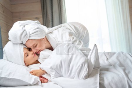 Téléchargez les photos : Jolie mère propre en serviette sur la tête embrassant sa douce petite fille en peignoir doux, les filles passent le week-end ensemble dans un centre de spa - en image libre de droit