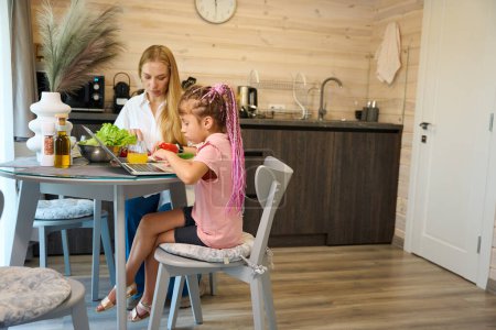 Photo for Schoolgirl doing her homework on laptop sitting on kitchen near focused on salad preparing mother, girls family spending time together - Royalty Free Image