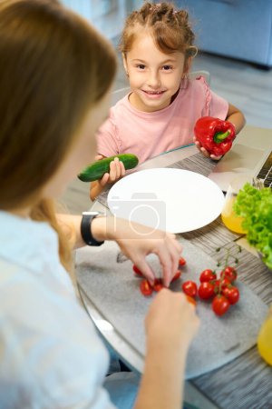 Foto de Dulce niña mirando a la cámara y ayudando a la madre sosteniendo pimiento rojo y pepino verde mientras su madre cortando tomates en ensalada - Imagen libre de derechos