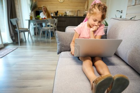Photo for Cute girl studying online, sitting on sofa with laptop and making test, doing her homework online, mom cooking and watching daughter from kitchen - Royalty Free Image