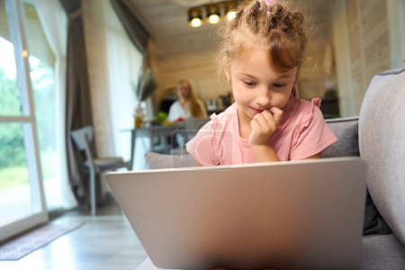Photo for Clever little girl looking at laptop display sitting on sofa in living room, girl doing her homework online or taking preparatory programming courses for children - Royalty Free Image