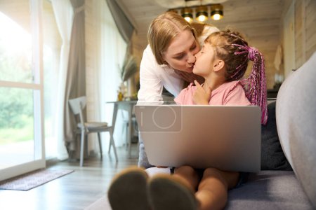 Téléchargez les photos : Mère embrasser sa petite fille qui prend des leçons en ligne assis sur le canapé avec ordinateur portable, enseignement à distance, cours d'étude supplémentaire - en image libre de droit