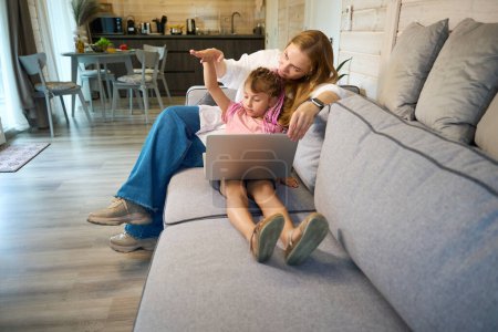 Téléchargez les photos : Mère attentionnée aidant sa petite écolière à faire des devoirs de mathématiques en ligne sur ordinateur portable, les filles comptant la hauteur et la distance avec les mains assises sur le canapé - en image libre de droit