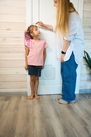 Photo for Mother measuring height of her curious daughter who standing near doorjamb and making mark with pencil, annual tradition, desire to grow up quickly - Royalty Free Image
