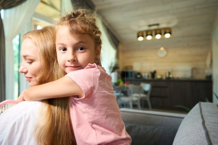 Photo for Pretty daughter hugging her attractive mother by shoulders, girls having fun sitting on sofa in living room, pleasure pastime, motherhood - Royalty Free Image
