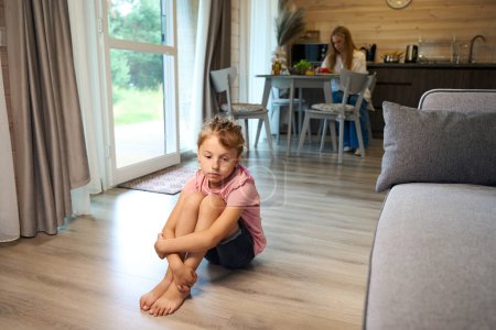 Photo for Upset little girl with braided hair lonely sitting on floor in living room while mother preparing dinner, has no friends and no mood for playing or helping to mother - Royalty Free Image