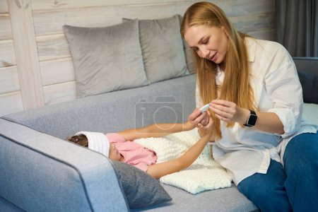 Téléchargez les photos : Mère vérifiant la température de la petite fille malade qui se trouve sur le canapé recouvert de plaid, fille souffrant de froid, se sentant malade - en image libre de droit