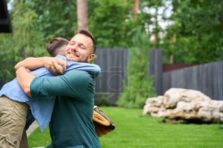 Foto de Feliz padre abraza a su hijo en el césped en casa, el niño tiene un guante de béisbol - Imagen libre de derechos