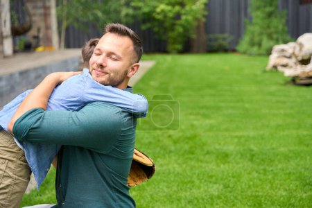 Foto de Sonriente padre abraza hijo en verde césped, chico tiene guante de béisbol - Imagen libre de derechos