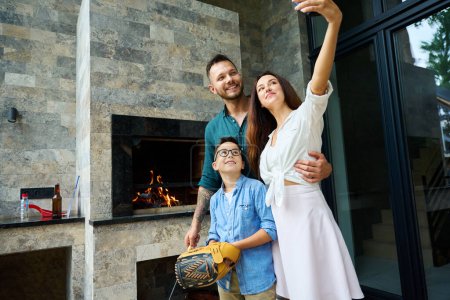 Photo for Woman takes a family selfie by the stove on the terrace, a man cooks food on the fire - Royalty Free Image