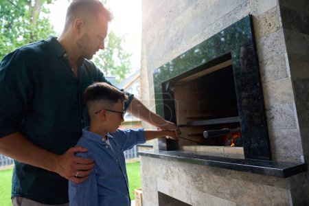 Téléchargez les photos : Papa et fils cuisinent sur un feu sur la terrasse, le poêle est décoré avec de la pierre décorative - en image libre de droit